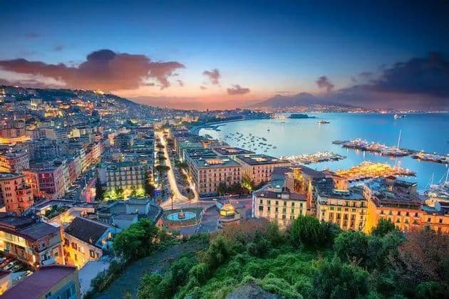 An elevated view of an illuminated coastal city curving around a bay at dusk, with boats in the harbor and mountains on the horizon.