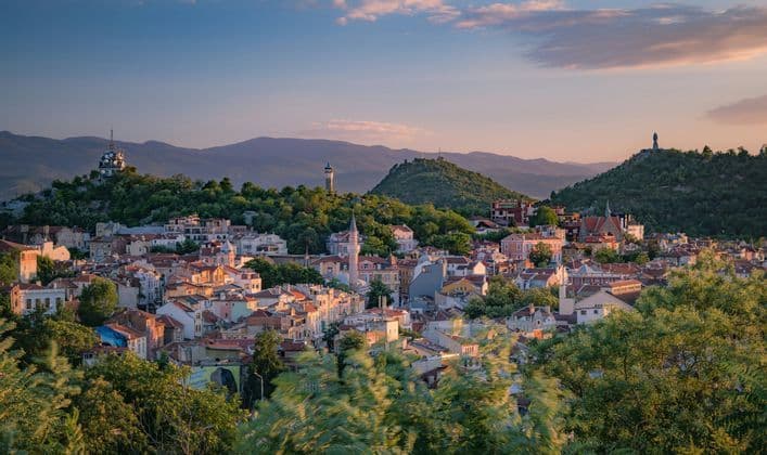 Une vue panoramique d'une ville colorée aux toits de tuiles, nichée entre des collines verdoyantes, au coucher de soleil doré.