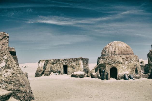 Mehrere sandfarbene Lehmziegelbauten, einer davon mit Kuppeldach, stehen in einer weiten Wüstenlandschaft unter blauem Himmel.