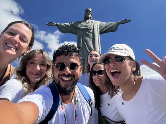 Un gruppo WeRoad sorride per un selfie davanti alla statua del Cristo Redentore sotto un cielo blu.