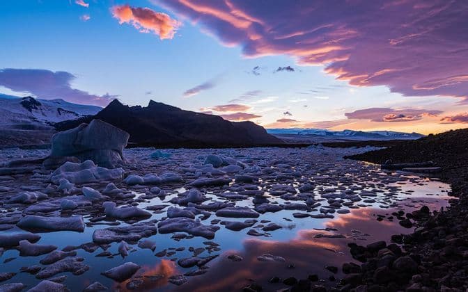 Iceberg e blocchi di ghiaccio galleggiano in una laguna glaciale al tramonto, riflettendo le nuvole viola e rosa nell'acqua calma.