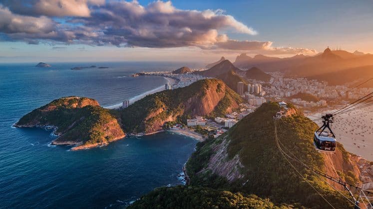 Una funivia scende su una città costiera con montagne verdi e una lunga spiaggia durante un tramonto dorato.