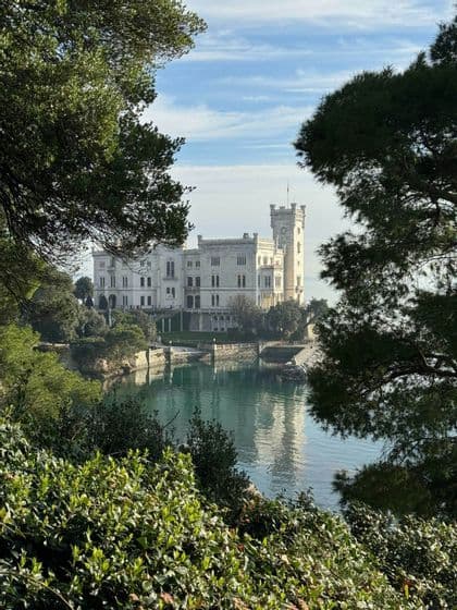 Un castello bianco con una torre si staglia sulla riva di acque calme, incorniciato da alberi e arbusti verdi.