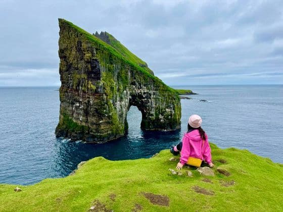 Una donna con una giacca rosa siede su una scogliera erbosa, guardando un grande faraglione coperto di muschio con un arco naturale nel mare.