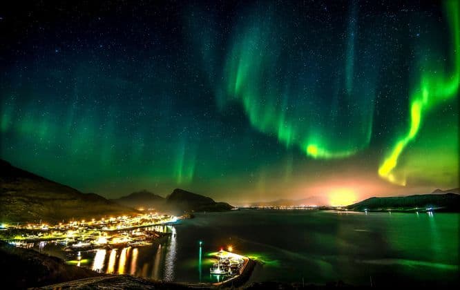 L'aurora boreale verde brilla nel cielo notturno stellato sopra una città costiera illuminata e un tranquillo specchio d'acqua.