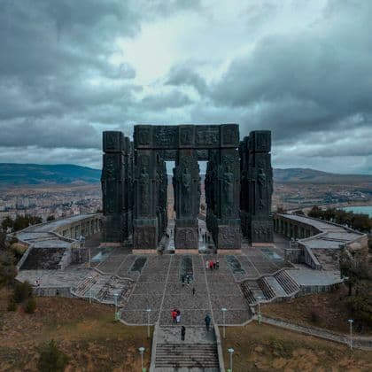 Una veduta aerea di un imponente monumento scolpito su una collina, con persone che camminano nella piazza sottostante sotto un cielo nuvoloso.