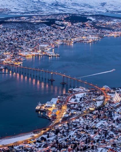 Eine Luftaufnahme einer schneebedeckten Küstenstadt in der Dämmerung, mit einer langen, beleuchteten Brücke, die über das Wasser zu verschneiten Bergen führt.