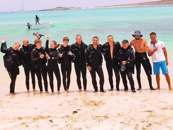 Un viaggio di gruppo WeRoad di subacquei in mute in posa per una foto su una spiaggia sabbiosa vicino ad acqua turchese.