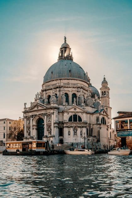 An ornate domed basilica stands on a canal waterfront, backlit by the sun, with boats in the foreground.