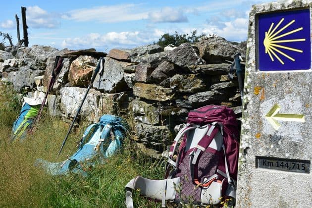 Zaini da trekking e bastoncini da trekking riposano nell'erba alta contro un muro di pietra vicino a un segnavia del Cammino di Santiago.