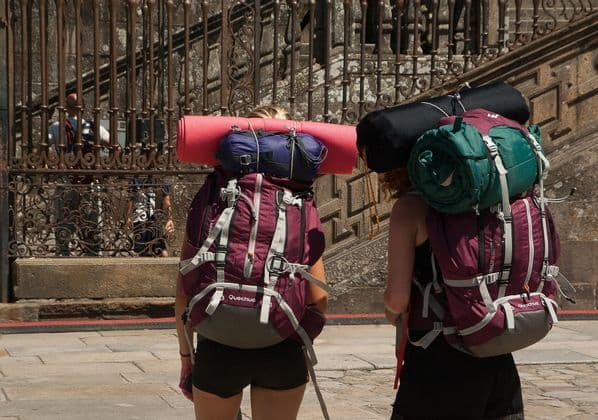 Le schiene di due persone di un viaggio di gruppo WeRoad con grandi zaini da trekking e sacchi a pelo fissati, davanti a un cancello di metallo decorato.