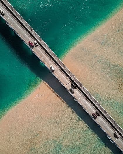 Una vista aerea dall'alto di auto in transito su un ponte che attraversa acqua turchese e un litorale sabbioso.