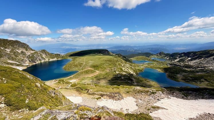 Une vue panoramique sur plusieurs lacs alpins bleus, disséminés dans une vallée de montagne verdoyante, avec des plaques de neige sous un ciel bleu nuageux.
