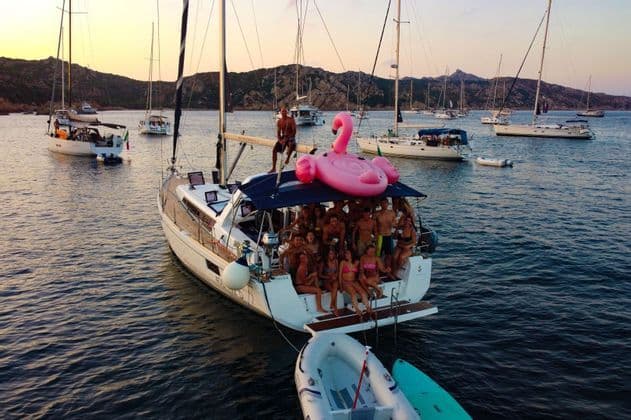 Un viaggio di gruppo WeRoad sorride per una foto su una barca a vela, con un grande fenicottero gonfiabile rosa, circondato da altre barche al tramonto.