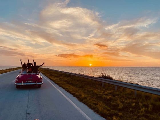 Un viaggio di gruppo WeRoad visto da dietro, alla guida di una decappottabile vintage rosa su una strada costiera durante un tramonto dorato.
