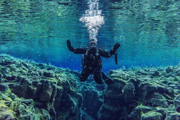 Una persona con equipo completo de buceo flota en agua clara de color turquesa, saludando con ambas manos sobre un cañón rocoso cubierto de algas.