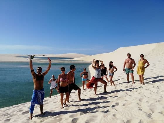 A WeRoad group trip posing on a white sand dune beside a turquoise lagoon under a clear sunny sky.