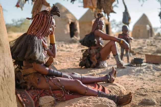 A woman in traditional attire with an intricate hairstyle sits on a rock outside a hut in a village.