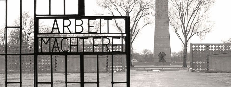 Una foto in bianco e nero di un cancello di ferro con la scritta 'Arbeit Macht Frei', con un monumento e alberi spogli sullo sfondo.