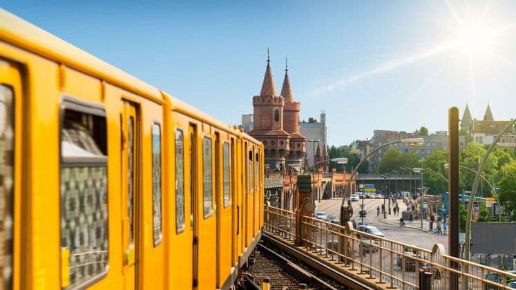 Un treno giallo viaggia su un binario sopraelevato attraverso un ponte, con un ponte storico della città e edifici sullo sfondo.