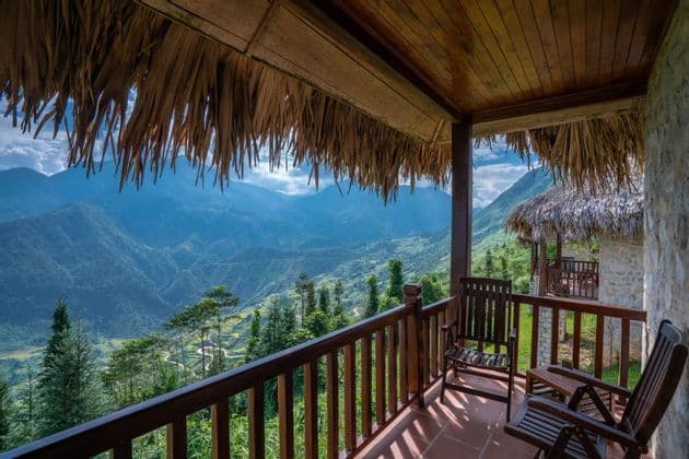 Une vue depuis un balcon au toit de chaume avec deux chaises en bois dominant une vaste vallée montagneuse et verdoyante par une journée ensoleillée.