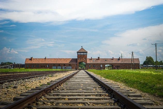 Una vista en contrapicado de vías de tren que conducen hacia un gran edificio de ladrillo con una torre de vigilancia central y una entrada arqueada.