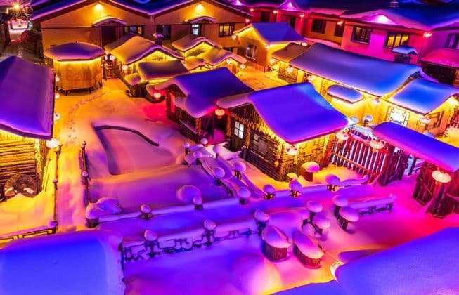Veduta aerea di un villaggio innevato di notte, con edifici illuminati da luci colorate gialle e viola.