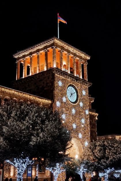 Une tour de l'horloge en pierre, illuminée de lumières festives et d'étoiles la nuit, avec le drapeau arménien flottant à son sommet.