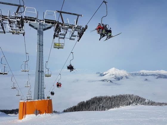Sciatori in seggiovia sopra un mare di nuvole, su una montagna innevata, con cime distanti visibili sotto un cielo sereno.