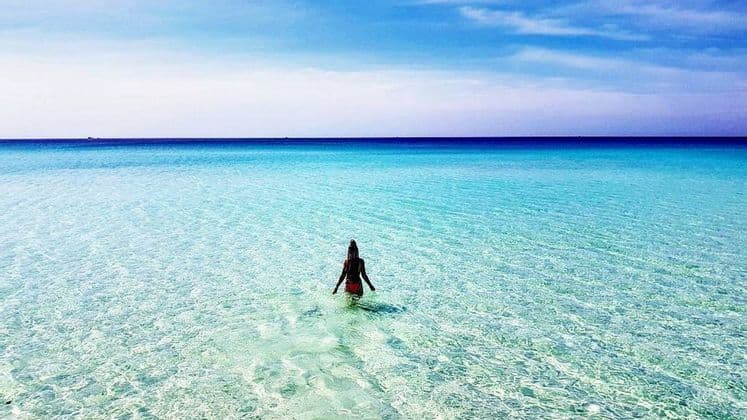 Una donna di spalle si addentra nel mare turchese e limpido sotto un cielo azzurro brillante.