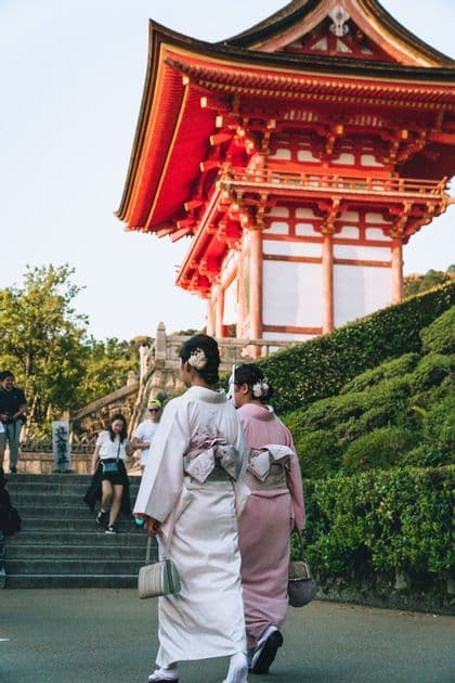 Zwei Frauen in traditionellen Kimonos gehen auf einem Pfad zu einer roten japanischen Pagode auf einem Hügel, von hinten gesehen.