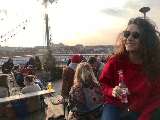 Una donna con un maglione rosso, partecipante a un viaggio di gruppo WeRoad, sorride mentre sorseggia un drink su un affollato rooftop bar al tramonto.