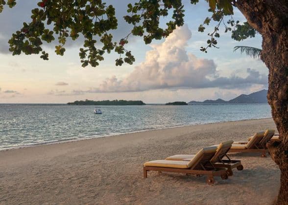 Des transats vides reposent sur une plage de sable face à la mer, au crépuscule, avec un bateau et des îles visibles au loin sous un ciel nuageux.