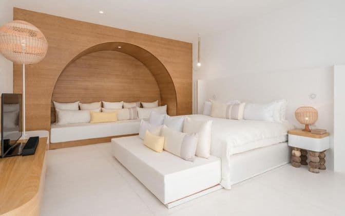 Una camera da letto bianca minimalista con un ampio letto e una parete ad arco in legno con un divano integrato, decorata con cuscini chiari.