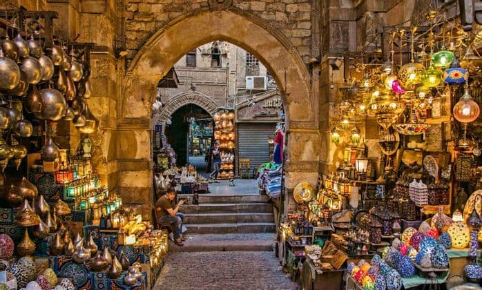 Une vue à travers une arcade de pierre vers une ruelle de marché animée, remplie d'étals vendant des lanternes colorées et illuminées et de l'artisanat.