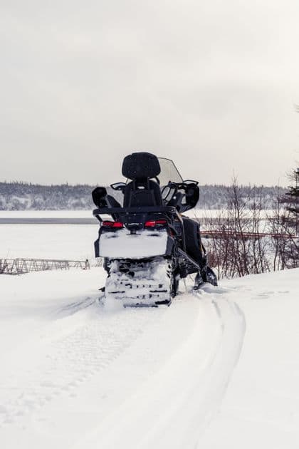 Una motoslitta nera è parcheggiata nella neve profonda, lasciando tracce dietro di sé, con un lago ghiacciato e alberi sullo sfondo.
