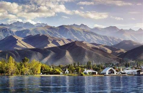 Un lago en calma se extiende frente a una orilla con árboles verdes y edificios blancos, con una vasta y ondulante cordillera de fondo bajo un cielo parcialmente nublado.