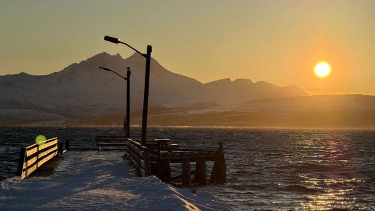 A snowy wooden pier extends into a body of water at sunset, with a backdrop of snow-covered mountains.