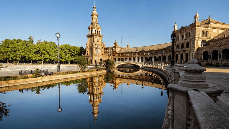 Un sontuoso edificio con un'alta torre si specchia nell'acqua calma di un canale che scorre sotto un ponticello in una giornata di sole.