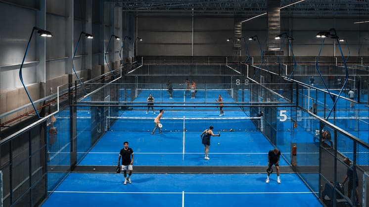 Una veduta dall'alto di persone che giocano a padel su diversi campi indoor con superfici blu brillanti, illuminati da grandi luci a soffitto.