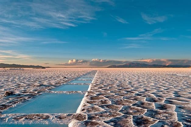 Saline poligonali con una fila di pozze d'acqua rettangolari che si estendono verso montagne lontane sotto un cielo blu.