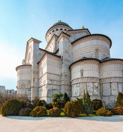 Una grande cattedrale in pietra bianca con cupola centrale e pareti decorate sorge in un giardino con arbusti verdi sotto un cielo azzurro e sereno.