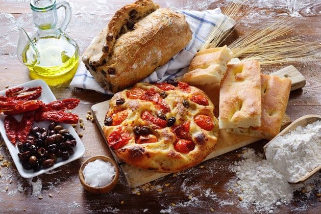 Un assortimento di pani italiani, inclusi focaccia e pane alle olive, con olio d'oliva, olive e pomodori su un tavolo di legno infarinato.