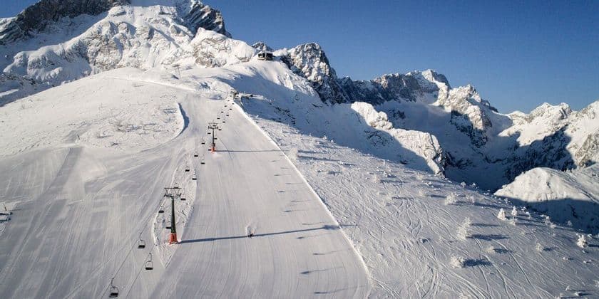 Ein einsamer Skifahrer fährt auf einer breiten, präparierten Skipiste einen weitläufigen, schneebedeckten Berg hinab, unter klarem Himmel und neben einem Sessellift.