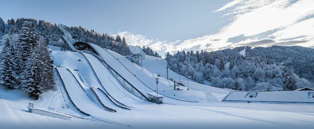 Eine große, schneebedeckte Skisprunganlage befindet sich an einem Berghang, umgeben von Kiefern, unter einem teils bewölkten Himmel.