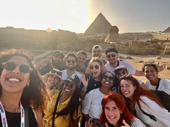 A WeRoad group trip takes a smiling selfie in front of the Great Pyramid and Sphinx under a golden sun.