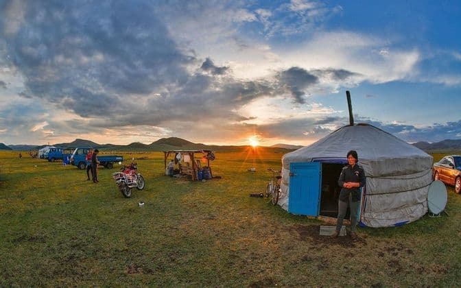 A yurt campsite on a wide grassy plain at sunset, with people, vehicles, and hills on the horizon.