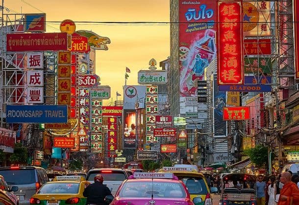 Una trafficata strada cittadina è piena di auto e taxi, affiancata da edifici ricoperti da innumerevoli insegne colorate sotto un cielo caldo e arancione.