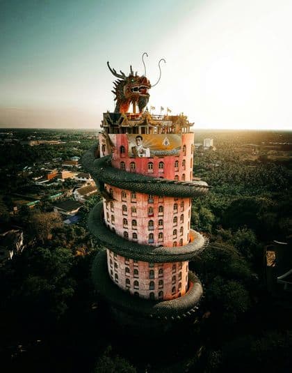 Una scultura di un drago gigante e scuro si avvolge attorno a un alto edificio cilindrico rosa, circondato da una foresta lussureggiante al tramonto.