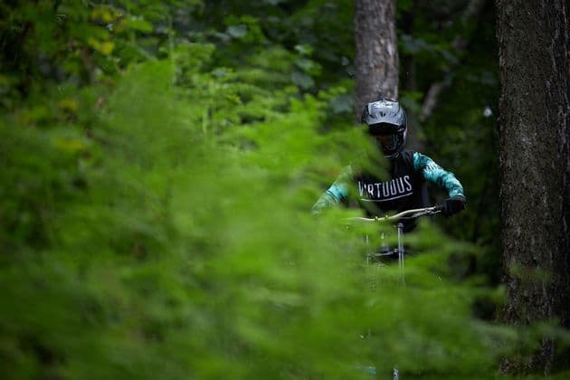 Un mountain biker con casco integrale e maglia attraversa una fitta foresta, visto da dietro il fogliame verde sfocato.